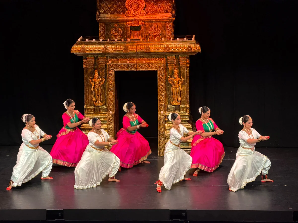 Seven dancers in pink and green or ivory costumes performing in front of an ornate gold stage backdrop.