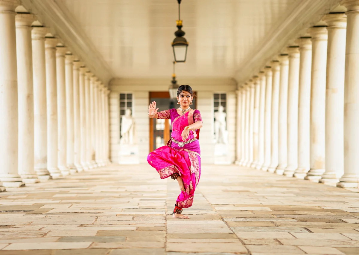 A solo dancer in a vivid pink costume with patterned gold borders, framed by a stone colonnade.