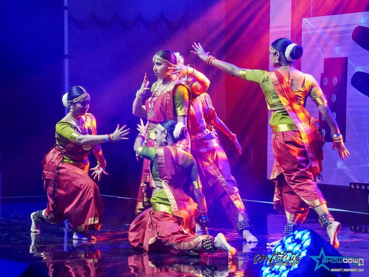A group of dancers in pink and green costumes with gold borders performing under colourful stage lights.